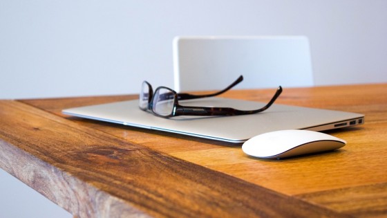 Macbook,glasses and a wireless mouse sat on a modern wooden desk