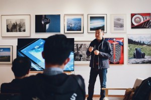Man standing up at the front of a hall next to a screen giving a presentation